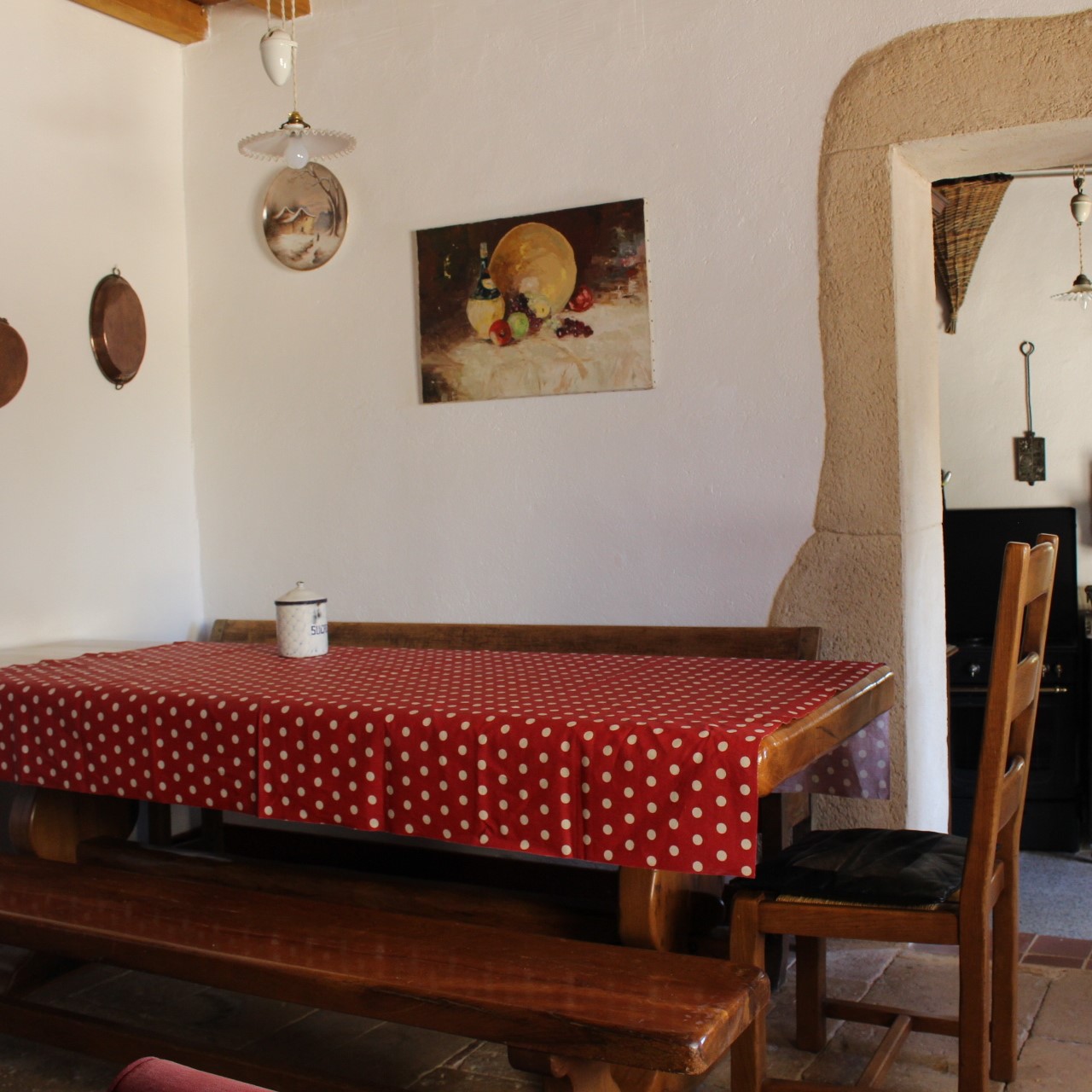 salon avec une table et une nappe, deux bancs, une chaise, une ouverture sur la cuisine à l'arrière